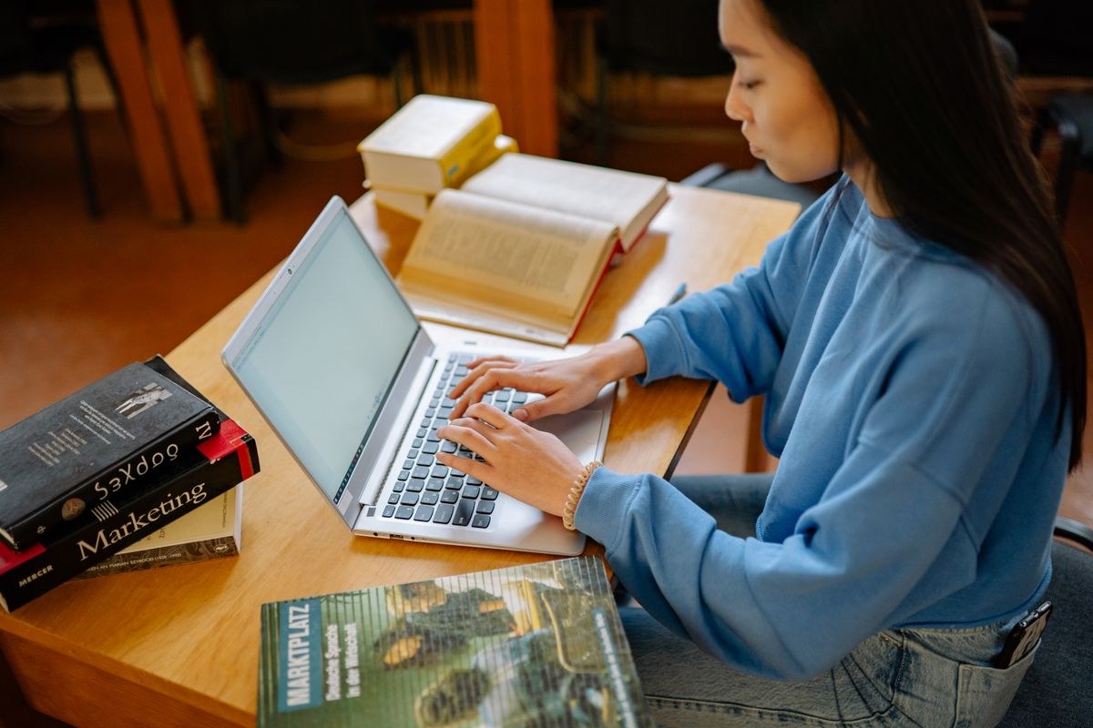Student typing on a laptop beside a stack of textbooks including a Marketing spine