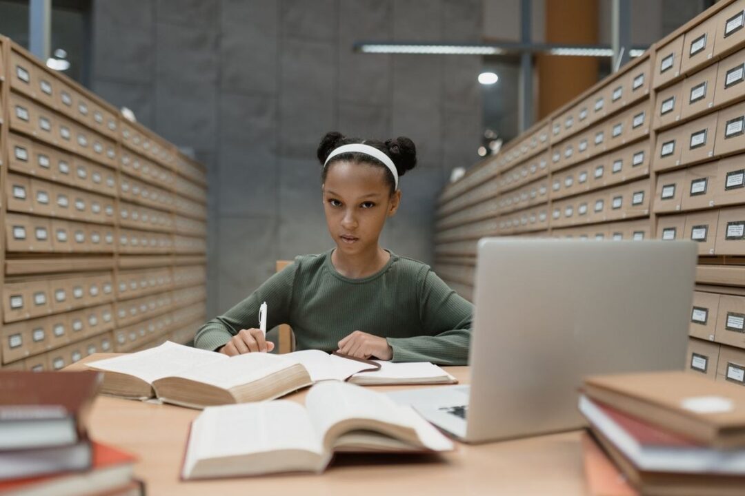 Student at a library desk with open textbooks, a laptop and a pen, looking toward the camera